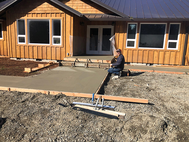 construction worker leveling a drying concrete pad outside, looking toward the building